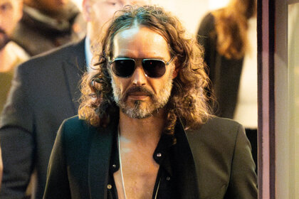 Russell Brand walking out of a building wearing sunglasses not smiling