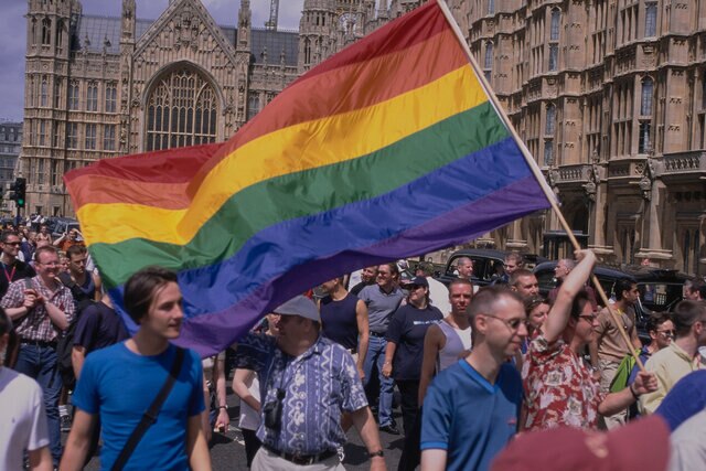 A large crowd of men attend a Pride event and wave a rainbow flag.